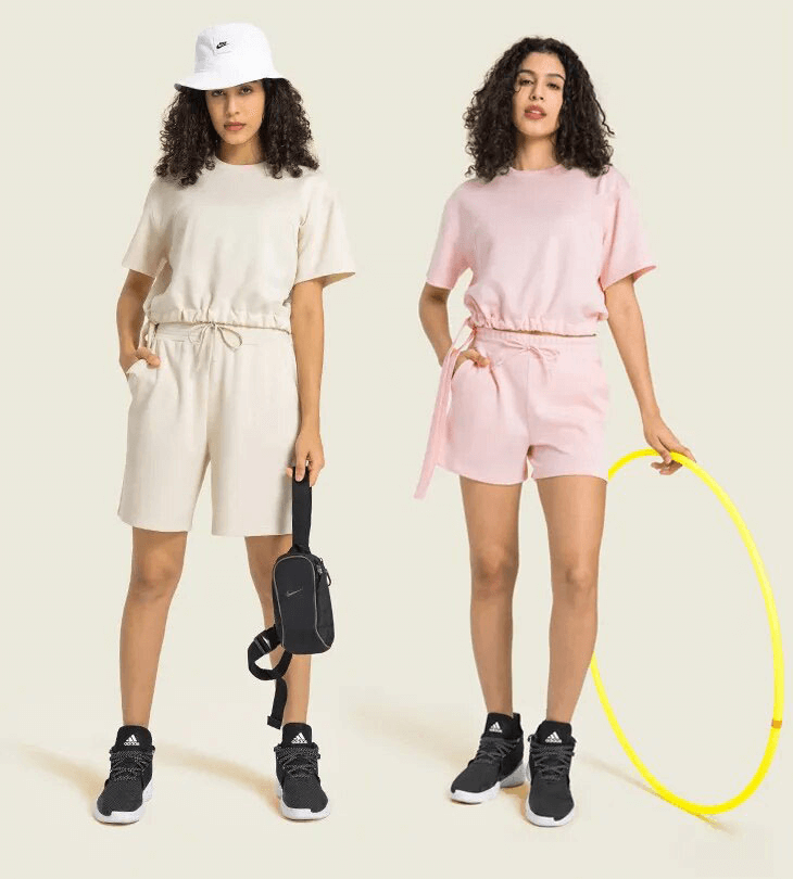 Two women wearing cream and pink coordinated sports sets with drawstring shorts, holding accessories, standing on beige background.